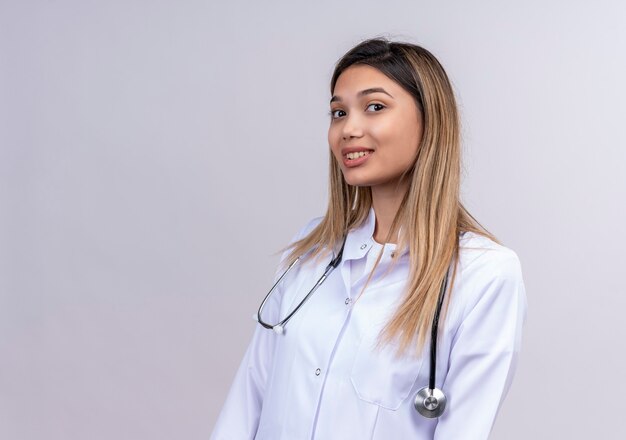 Belle jeune femme médecin portant blouse blanche avec stéthoscope à la recherche avec un sourire confiant sur le visage