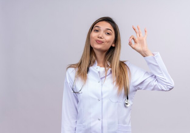 Belle jeune femme médecin portant blouse blanche avec stéthoscope souriant amical faisant signe ok