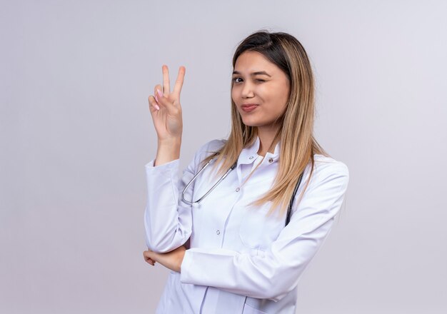 Belle jeune femme médecin portant blouse blanche avec stéthoscope souriant et clignotant montrant le signe de la victoire