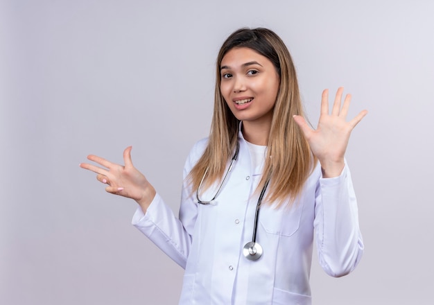 Belle jeune femme médecin portant blouse blanche avec stéthoscope souriant montrant confiant et pointant vers le haut avec les doigts numéro huit