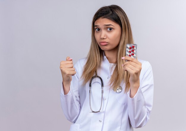 Belle jeune femme médecin portant blouse blanche avec stéthoscope tenant blister avec pilules serrant le poing à mécontent