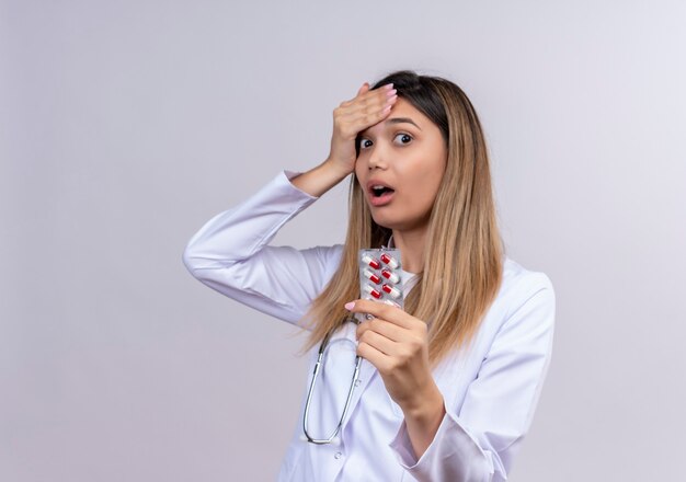 Belle jeune femme médecin portant blouse blanche avec stéthoscope tenant blister avec des pilules à la surprise et surpris