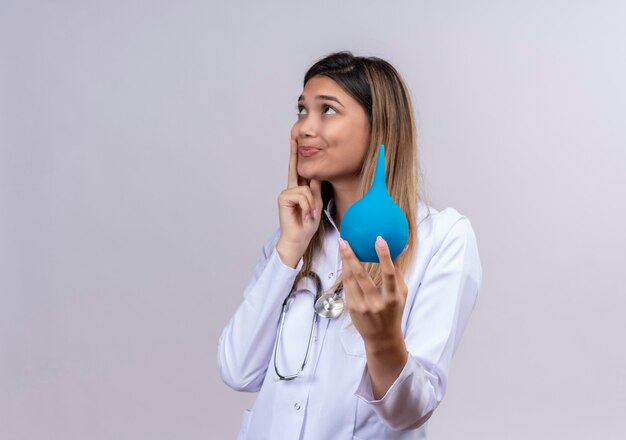 Belle jeune femme médecin portant blouse blanche avec stéthoscope tenant un lavement à la recherche avec une expression pensive sur le visage