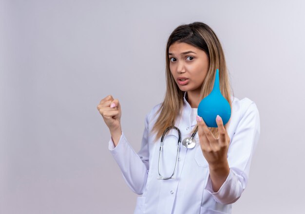 Belle jeune femme médecin portant blouse blanche avec stéthoscope tenant un lavement serrant le poing comme un gagnant