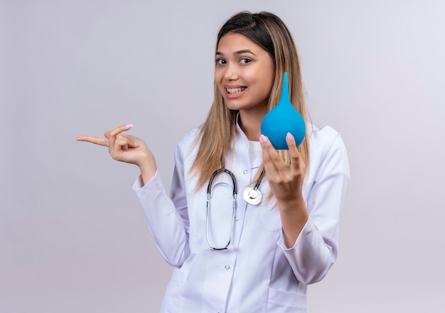 Belle jeune femme médecin portant un blouse blanche avec stéthoscope tenant un lavement souriant joyeusement pointant avec le doigt sur le côté