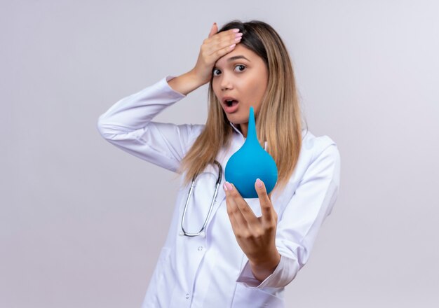 Belle jeune femme médecin portant blouse blanche avec stéthoscope tenant un lavement à la surprise et étonné de toucher sa tête