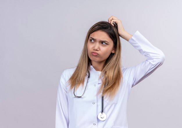 Belle jeune femme médecin vêtu d'un blouse blanche avec stéthoscope à la confusion avec le visage fronçant se gratter la tête ayant des doutes