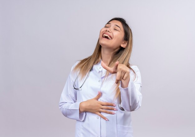 Belle jeune femme médecin vêtu d'un blouse blanche avec stéthoscope plaisantant avec quelqu'un en riant pointant avec l'index