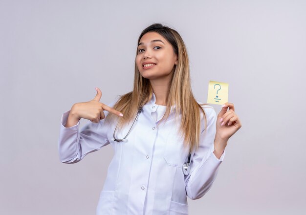 Belle jeune femme médecin vêtu d'un blouse blanche avec stéthoscope tenant un papier de rappel avec point d'interrogation pointant avec l'index pour positif et heureux