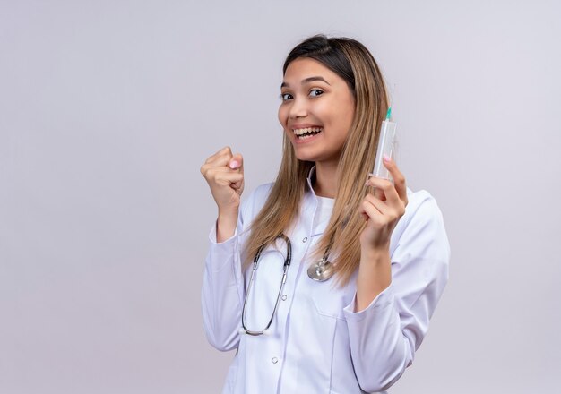 Belle Jeune Femme Médecin Vêtu D'un Blouse Blanche Avec Stéthoscope Tenant La Seringue Serrant Le Poing Heureux Et Sortit En Se Réjouissant De Son Succès