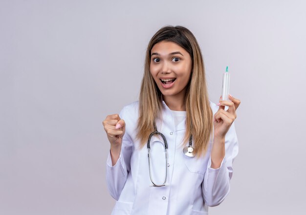 Belle jeune femme médecin vêtu d'un blouse blanche avec stéthoscope tenant la seringue serrant le poing heureux et sortit en se réjouissant de son succès