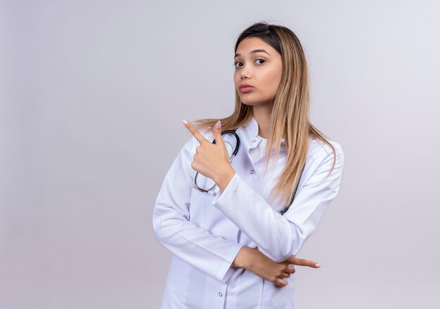 Belle jeune femme médecin vêtu d'un manteau blanc avec stéthoscope à la confiance en pointant avec l'index à quelque chose derrière