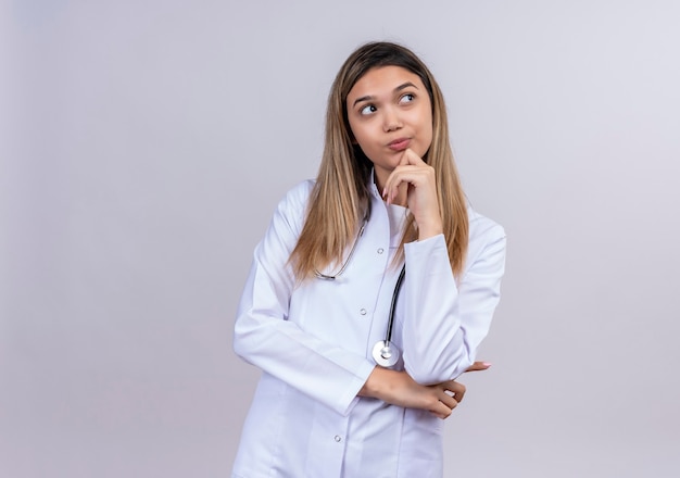 Belle jeune femme médecin vêtu d'un manteau blanc avec stéthoscope à côté avec la main sur le menton avec une expression pensive sur le visage