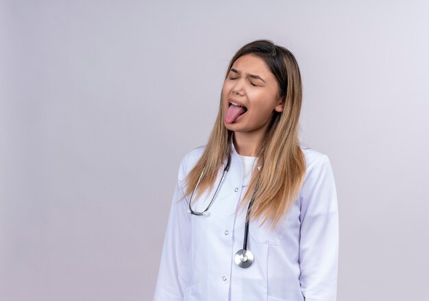 Belle jeune femme médecin vêtu d'un manteau blanc avec stéthoscope ennuyé et dérangé qui sort la langue