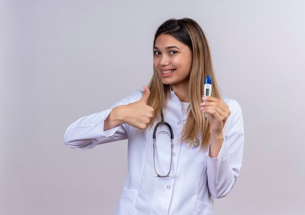 Belle jeune femme médecin vêtu d'un manteau blanc avec stéthoscope tenant un thermomètre numérique souriant avec visage heureux montrant les pouces vers le haut