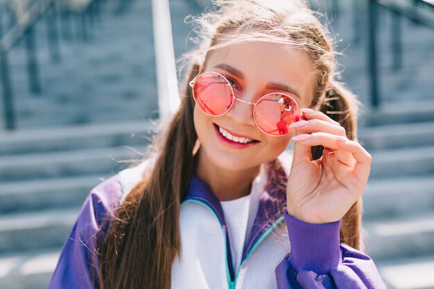 Belle jeune femme à la mode dans des vêtements élégants portant des lunettes de soleil roses et souriant