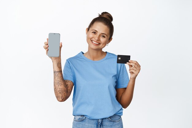 Belle jeune femme montrant un écran de téléphone vide avec carte de crédit, interface d'application, souriant sur blanc.