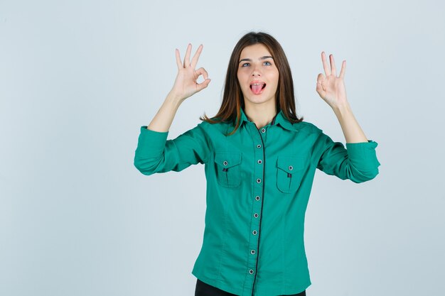 Belle jeune femme montrant un geste ok tout en sortant la langue en chemise verte et à la drôle, vue de face.