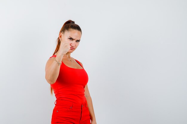 Belle jeune femme montrant le poing fermé en débardeur rouge, pantalon et à la méchante, vue de face.