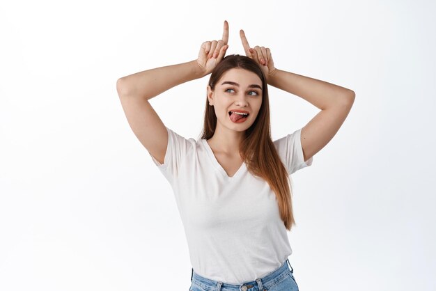 Belle jeune femme montre le geste de la langue et des cornes de taureau en regardant de côté l'espace de copie avec le signe du logo de lecture de texte promotionnel debout en t-shirt sur fond blanc