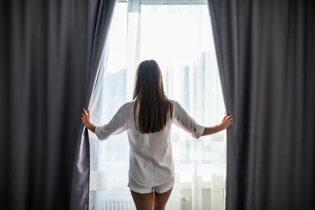 Belle jeune femme ouvrant des rideaux et regardant par la fenêtre