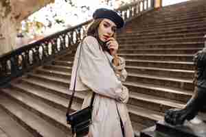 Photo gratuite belle jeune femme parisienne aux cheveux bruns en béret élégant, trench-coat beige et sac noir, debout sur de vieux escaliers et posant avec sensibilité à l'extérieur