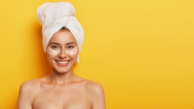 belle jeune femme avec une peau douce et saine sous les yeux, porte une serviette sur la tête, applique des patchs pour réduire les ridules, se tient nue contre le mur jaune. traitement de Spa