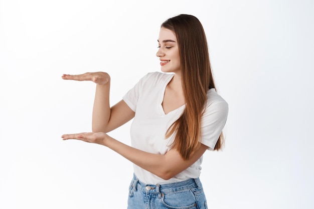 Belle jeune femme avec une peau hydratée saine tient un espace vide dans le geste de la boîte montrant l'espace de copie dans ses mains debout sur fond blanc afficher le logo ou l'article de la société