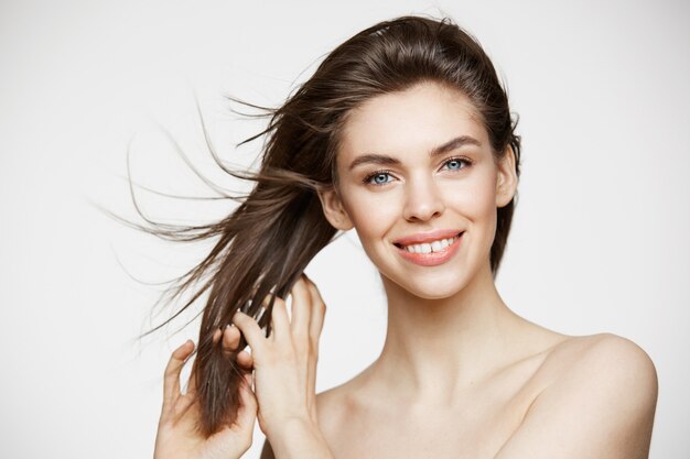 Belle jeune femme avec une peau parfaitement propre, souriant, toucher les cheveux sur le mur blanc. Traitement facial.