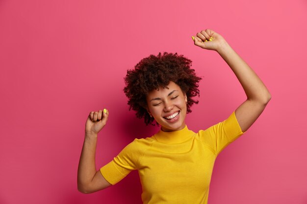 Belle jeune femme à la peau sombre se sent détendue et soulagée, danse sans soucis, lève les bras en l'air, sourit positivement, habillée avec désinvolture, pose contre le mur rose. Concept de mode de vie heureux