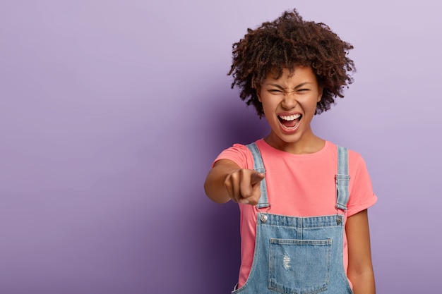 Photo gratuite belle jeune femme à la peau sombre vous accuse de faire quelque chose de mal, pointe l'index directement sur la caméra, crie négativement, garde les yeux fermés, sourit narquois, blâme quelqu'un, pose à l'intérieur