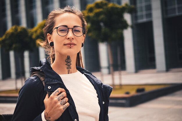 Belle jeune femme avec piercing, lunettes et tatouage sur son cou pose pour le photographe dans la rue.