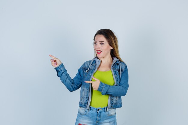 Belle jeune femme pointant vers la gauche, regardant de côté en tenue de denim et semblant joyeuse. vue de face.