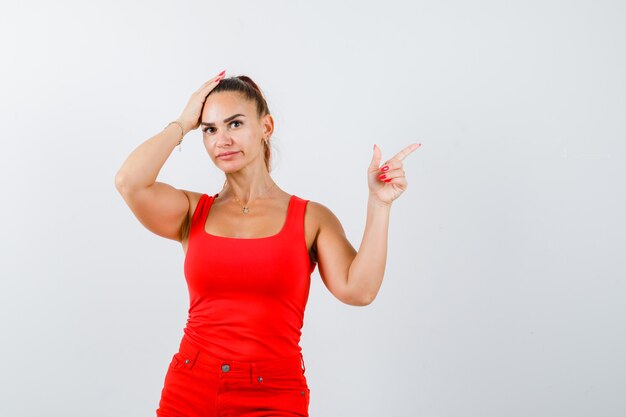 Belle jeune femme pointant vers le haut, tenant la main sur la tête en débardeur rouge, pantalon et à la vue troublée, de face.