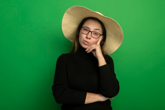 Belle jeune femme portant un chapeau d'été dans un col roulé noir et des lunettes à l'avant avec une expression sceptique debout sur un mur vert