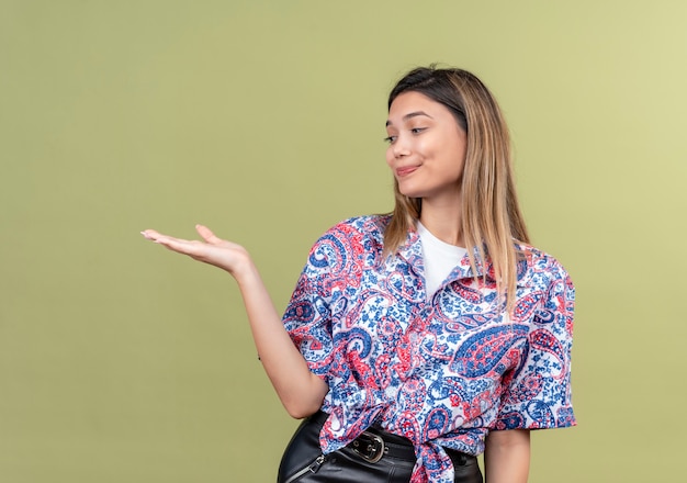 Photo gratuite une belle jeune femme portant une chemise imprimée paisley en levant la main et en la regardant sur un mur vert