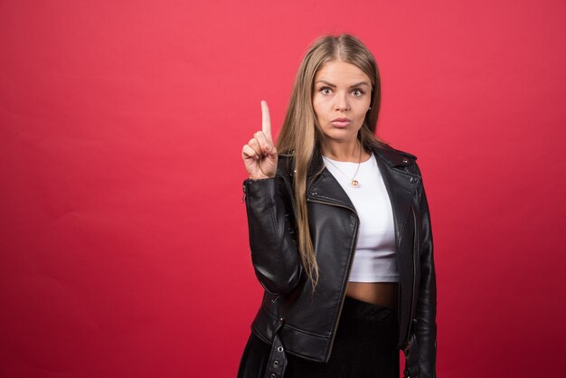 Belle jeune femme portant une veste en cuir debout sur un mur rouge et pointant le doigt vers le haut