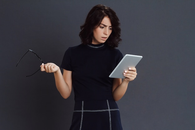 Belle jeune femme posant sur un mur gris tenant une tablette et des lunettes dans les mains