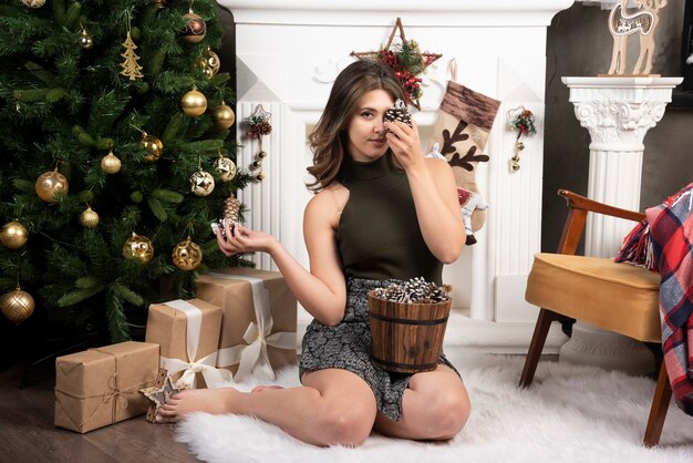 Belle jeune femme posant avec panier de pommes de pin près de l'arbre de Noël