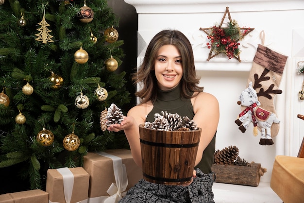 Belle jeune femme posant avec panier de pommes de pin près de l'arbre de Noël