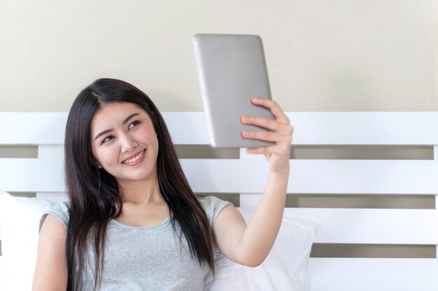 belle jeune femme prenant une photo elle-même, elle sourit avec bonheur et selfie par grand smartphone sur le lit à la maison