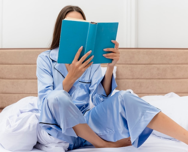 Belle Jeune Femme En Pyjama Bleu Assis Sur Le Lit Avec Livre De Lecture Livre Dans L'intérieur De La Chambre Sur Fond Clair