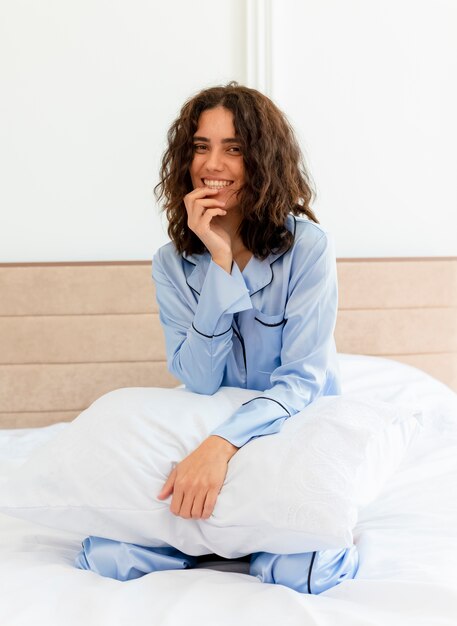 Belle jeune femme en pyjama bleu assis sur le lit avec oreiller au repos profitant du week-end heureux et positif souriant gaiement à l'intérieur de la chambre sur fond clair