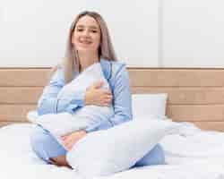Photo gratuite belle jeune femme en pyjama bleu assis sur le lit avec oreiller lookign à la caméra souriant avec un visage heureux à l'intérieur de la chambre sur fond clair