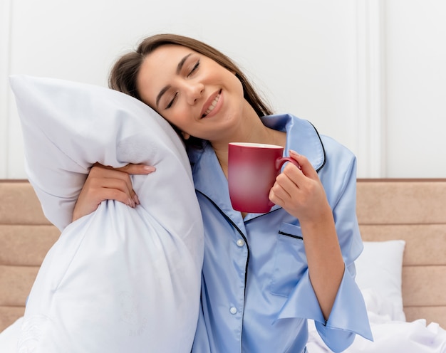 Belle jeune femme en pyjama bleu assis sur le lit avec oreiller et tasse de café, ressentir des émotions positives avec les yeux fermés à l'intérieur de la chambre sur fond clair