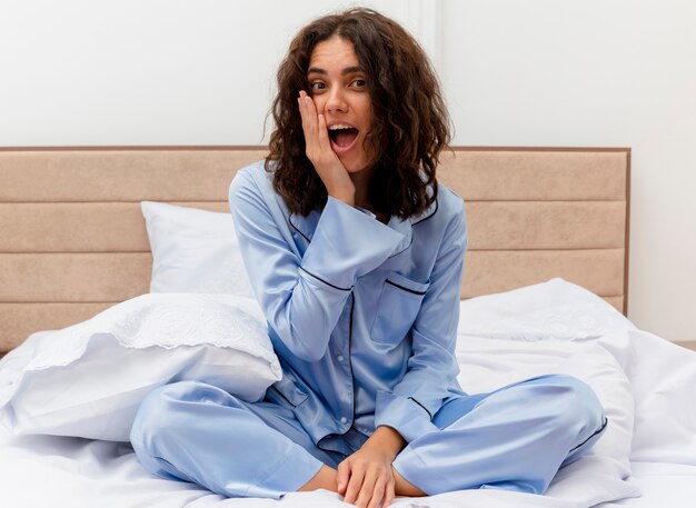 Belle jeune femme en pyjama bleu assis sur le lit en regardant la caméra étonné et surpris dans l'intérieur de la chambre sur fond clair