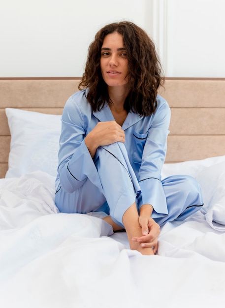 Belle jeune femme en pyjama bleu assis sur le lit en regardant la caméra avec le sourire sur le visage à l'intérieur de la chambre sur fond clair