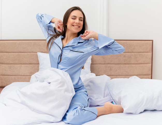 Belle jeune femme en pyjama bleu assis sur le lit se réveiller s'étendant les mains profitant de l'heure du matin dans l'intérieur de la chambre sur fond clair