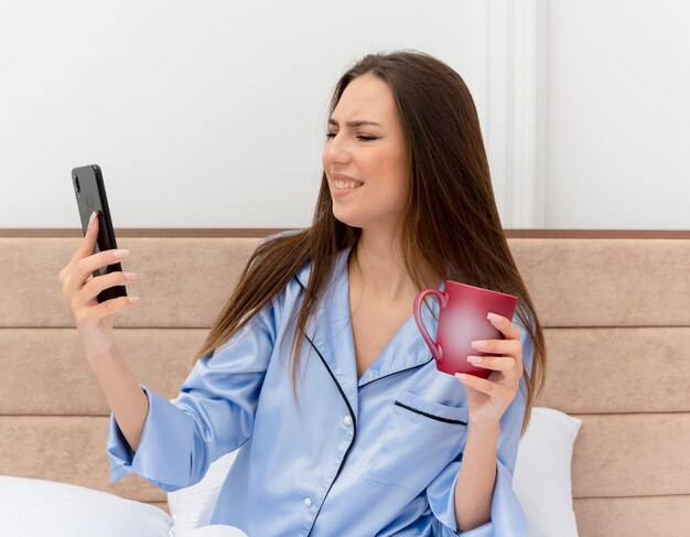 Belle jeune femme en pyjama bleu assis sur le lit avec une tasse de café à l'aide de smartphone à la mécontentement à l'intérieur de la chambre sur fond clair