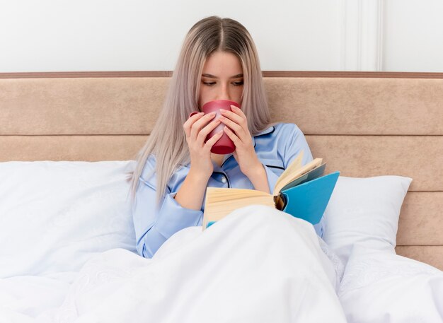 Belle jeune femme en pyjama bleu assis sur le lit avec tasse de café livre de lecture à l'intérieur de la chambre sur fond clair